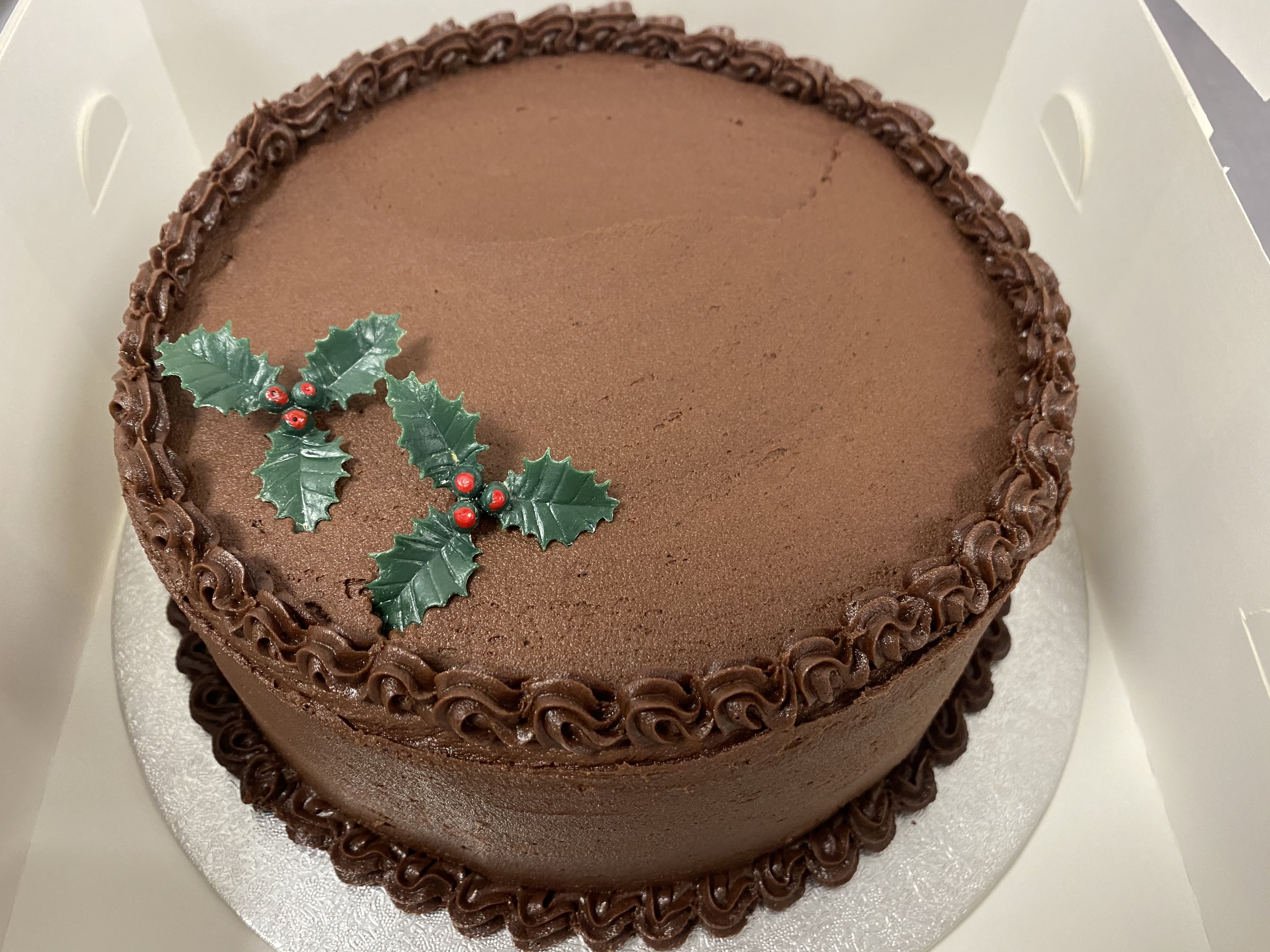 A round chocolate cake, a decadent dessert, is adorned with a border of chocolate frosting along the top and bottom edges. A small decoration of plastic holly leaves and red berries graces the top. The cake sits on a silver board inside a white box.